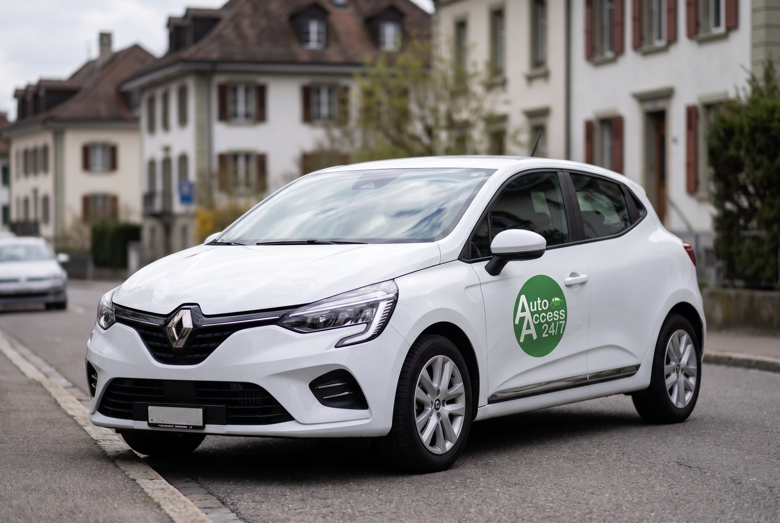 Citadine Renault Clio blanche au logo AutoAccess sur la portière, stationnée dans une rue résidentielle